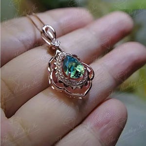 Puede incluir: Colgante de oro rosa con una piedra preciosa verde-azulada en forma de pera. La piedra est&aacute; rodeada de peque&ntilde;as piedras transparentes y engastada en un marco floral. El collar se muestra sobre un fondo borroso.