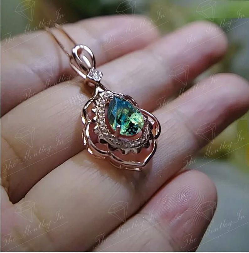 Puede incluir: Colgante de oro rosa con una piedra preciosa verde-azulada en forma de pera. La piedra est&aacute; rodeada de peque&ntilde;as piedras transparentes y engastada en un marco floral. El collar se muestra sobre un fondo borroso.