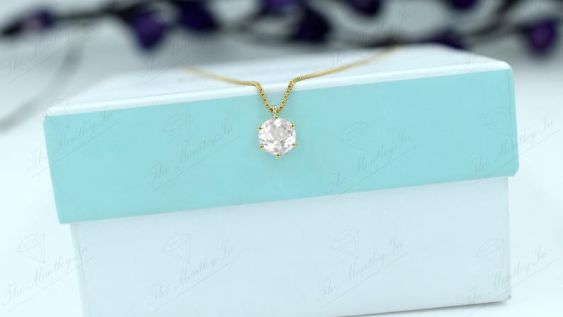 Pu&ograve; includere: Una collana d'oro con un pendente di pietra preziosa rosa chiaro, appoggiata su una scatola azzurra e bianca. Il pendente ha una forma esagonale. La collana &egrave; delicata ed elegante.