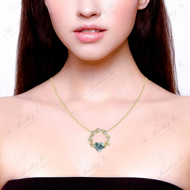 May include: A gold chain necklace with a pendant featuring a heart-shaped green gemstone surrounded by a wreath of leaves and small clear gemstones.