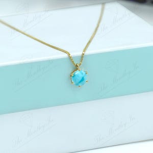 Op de afbeelding: Een gouden ketting met een blauwe edelsteen hanger. De hanger is een ronde, cabochon geslepen steen met een swirl-patroon. De ketting wordt gepresenteerd in een witte geschenkdoos met een lichtblauwe deksel.
