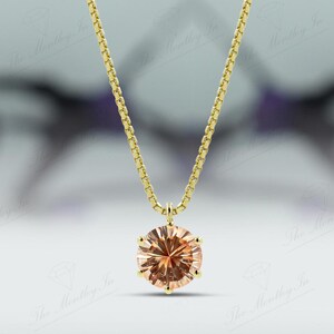 May include: A gold necklace with a round, peach-colored gemstone pendant. The pendant is the focal point, suspended from a delicate gold chain. The background is blurred, emphasizing the jewelry.
