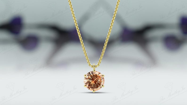 May include: A gold necklace with a round, peach-colored gemstone pendant. The pendant is the focal point, suspended from a delicate gold chain. The background is blurred, emphasizing the jewelry.