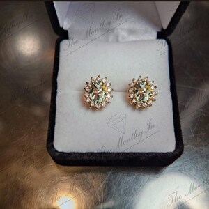 May include: A pair of gold earrings with oval-shaped green gemstones surrounded by smaller clear gemstones.