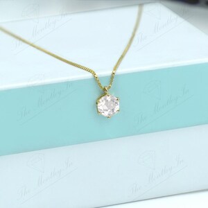 Pu&ograve; includere: Una collana color oro con un pendente rotondo in pietra preziosa rosa chiaro. La collana &egrave; esposta su una scatola bicolore con la parte superiore azzurra e la base bianca. La scatola &egrave; chiusa e sembra essere di cartone.