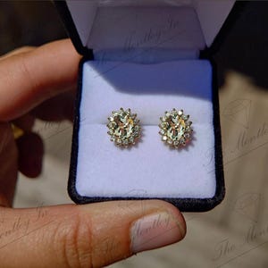 May include: A pair of oval-shaped earrings with a gold setting and a halo of smaller diamonds surrounding a larger center stone. The earrings are displayed in a white velvet box.