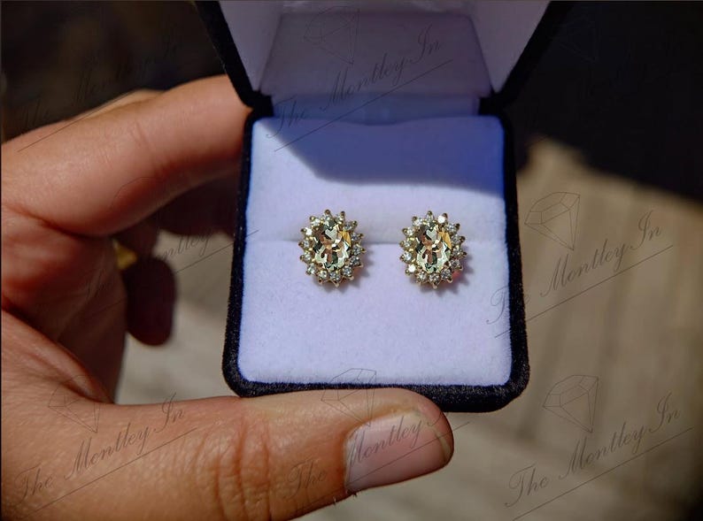 May include: A pair of oval-shaped earrings with a gold setting and a halo of smaller diamonds surrounding a larger center stone. The earrings are displayed in a white velvet box.