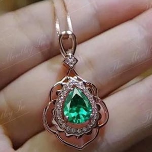 Puede incluir: Un collar con colgante de oro rosa con una piedra preciosa verde esmeralda en forma de pera, rodeada por un halo de pequeñas piedras transparentes. El colgante tiene un diseño calado ornamentado y cuelga de una delicada cadena.