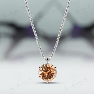 May include: A silver necklace with a round, light orange gemstone pendant. The pendant is the focal point, suspended from a delicate chain. The background is blurred, emphasizing the jewelry.