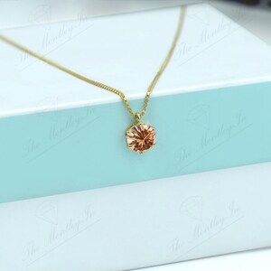May include: A gold-colored necklace with a round, peach-colored pendant. The necklace is displayed on a white and light blue gift box. The pendant has a faceted design, catching the light.