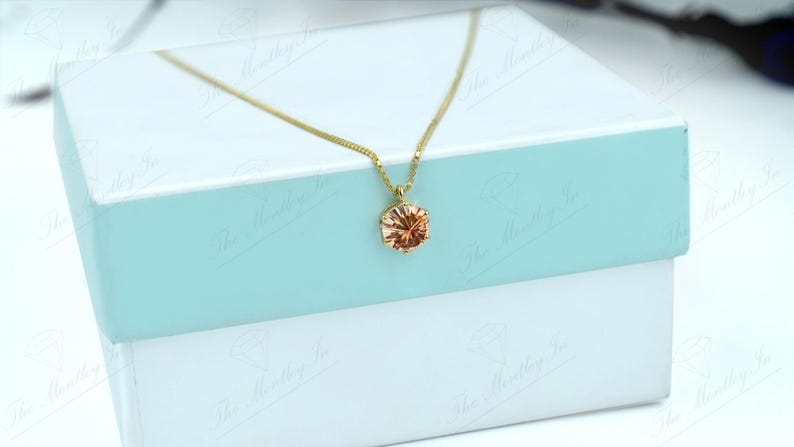 May include: A gold-colored necklace with a round, peach-colored pendant. The necklace is displayed on a white and light blue gift box. The pendant has a faceted design, catching the light.