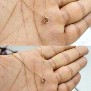 May include: A delicate gold necklace featuring a single, round, peach-colored gemstone pendant. The pendant is set in a simple, classic design, and the chain is a fine, gold link. The necklace is displayed on a hand.