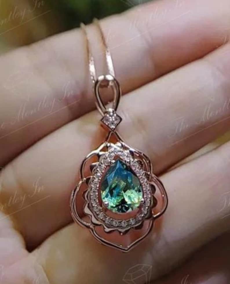 Puede incluir: Collar con colgante de oro rosa con una piedra preciosa en forma de pera, de color verde y azul. La piedra est&aacute; rodeada de peque&ntilde;os cristales transparentes y engastada en un marco calado decorativo. La cadena del collar es delicada.