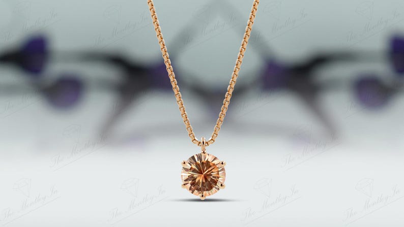 May include: A gold-colored necklace with a round, light brown gemstone pendant. The pendant is faceted, catching the light. The chain is a delicate box chain style. The background is blurred, with hints of purple and white.
