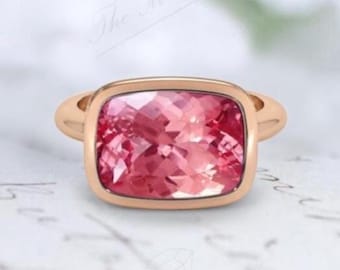 May include: A rose gold ring featuring a large, faceted, rectangular pink gemstone. The gemstone is set in a bezel setting, and the band has a smooth, rounded design. The ring is displayed on a white surface.