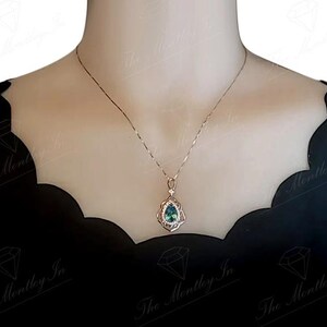 Puede incluir: Un delicado collar con un colgante en forma de l&aacute;grima. El colgante presenta una piedra preciosa de color verde azulado rodeada por un engaste de plata detallado y ornamentado. El collar se muestra sobre un fondo oscuro, resaltando el dise&ntilde;o de la joya.