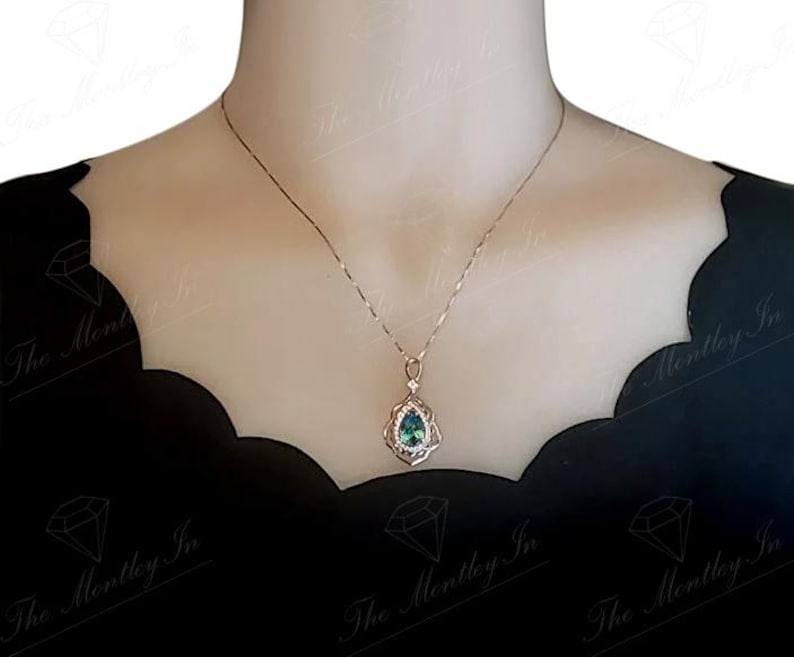 Puede incluir: Un delicado collar con un colgante en forma de l&aacute;grima. El colgante presenta una piedra preciosa de color verde azulado rodeada por un engaste de plata detallado y ornamentado. El collar se muestra sobre un fondo oscuro, resaltando el dise&ntilde;o de la joya.