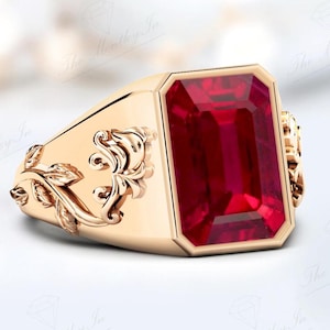 Puede incluir: Un anillo dorado con una gran gema rectangular de color rojo intenso. El anillo presenta detalles florales ornamentados en los laterales y un acabado pulido. La gema está engastada en un bisel.