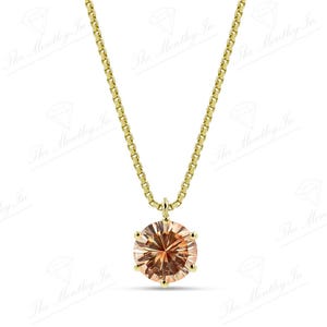 May include: A gold-colored necklace featuring a round, peach-colored gemstone pendant. The pendant is set in a gold-colored metal and hangs from a delicate gold-colored chain. The necklace is displayed against a white background.