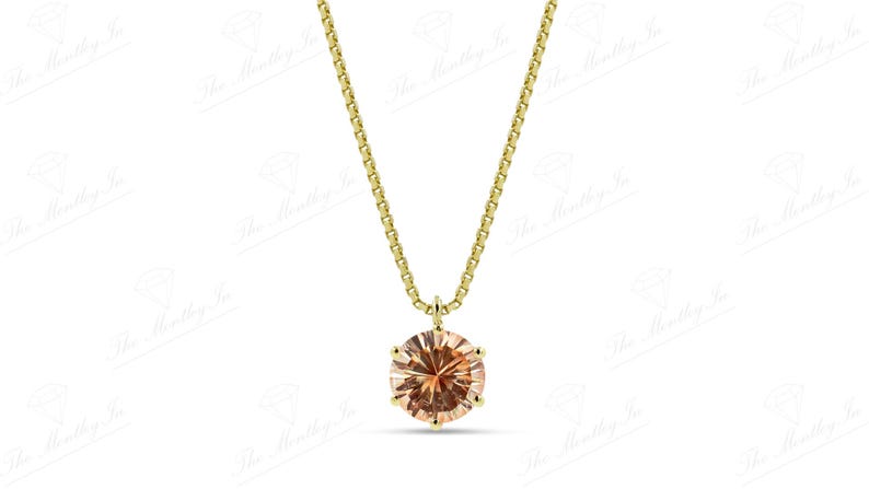 May include: A gold-colored necklace featuring a round, peach-colored gemstone pendant. The pendant is set in a gold-colored metal and hangs from a delicate gold-colored chain. The necklace is displayed against a white background.