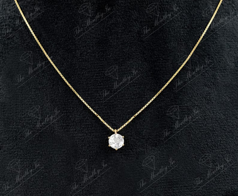 Pu&ograve; includere: Una delicata collana d'oro con un singolo pendente di pietra preziosa trasparente e rotonda. Il pendente &egrave; incastonato in una montatura a sei punte e pende da una sottile catena d'oro. Lo sfondo &egrave; nero.
