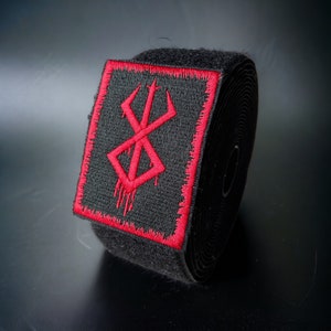 Könnte beinhalten: Schwarzer Klettverschluss mit einem roten gestickten Symbol. Das Symbol ist ein stilisiertes 'X' mit einem tropfenden Bluteffekt.