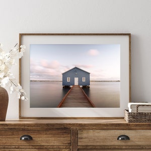 May include: A framed photograph of a blue boathouse at the end of a wooden pier extending into calm water. The sky is a soft pink and blue. The photograph is displayed on a wooden dresser with a vase of white flowers.