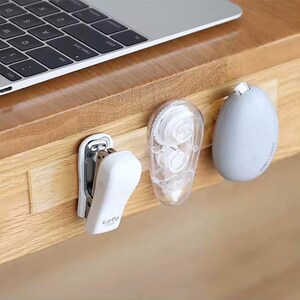 May include: A white tape dispenser, a white stapler labelled "EZFO", and a grey eraser are attached to a clear adhesive strip on a wooden surface.