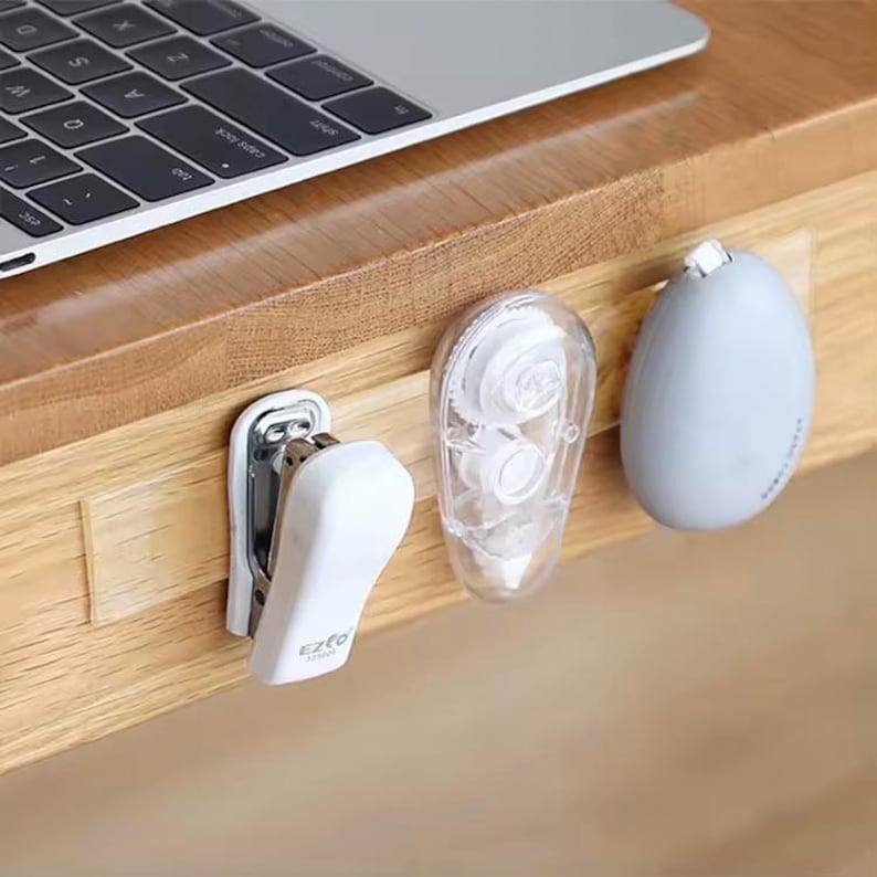 May include: A white tape dispenser, a white stapler labelled "EZFO", and a grey eraser are attached to a clear adhesive strip on a wooden surface.