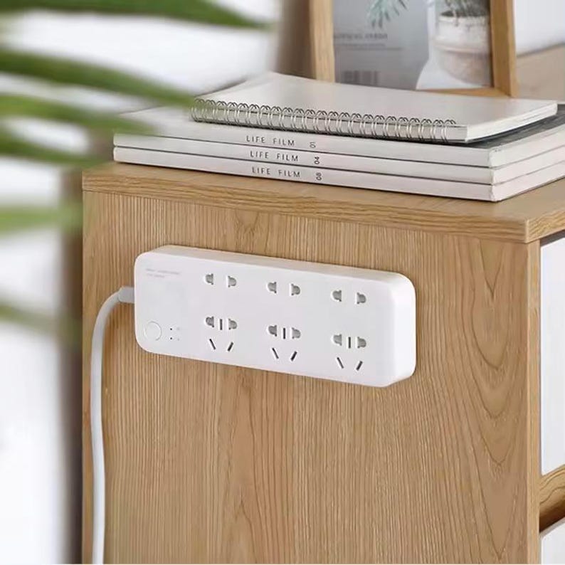 May include: White power strip with six sockets mounted on a wooden surface. The power strip has a white cord and a small white button on the side.