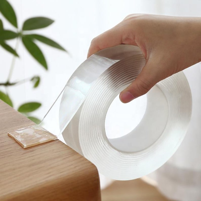 May include: A roll of clear, transparent adhesive tape. The tape is being unrolled from a dispenser and is being held in a hand.