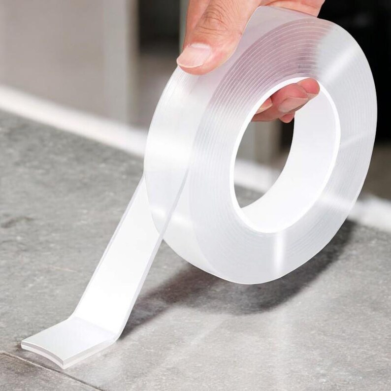 May include: A roll of clear, transparent adhesive tape. The tape is being unrolled and is lying on a white surface.