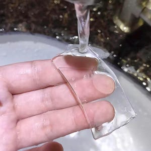 May include: A clear, flexible sheet of plastic being rinsed under a stream of water. The sheet is being held in a hand.