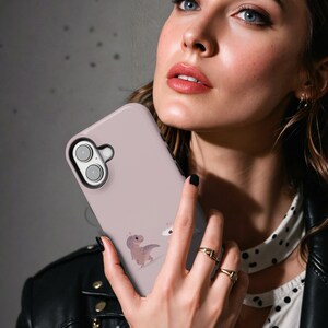 Puede incluir: Funda de tel&eacute;fono rosa claro con un dise&ntilde;o de dinosaurio y cupcake de dibujos animados. La funda tiene un recorte negro para la c&aacute;mara. El fondo es gris borroso. La funda est&aacute; siendo sostenida.