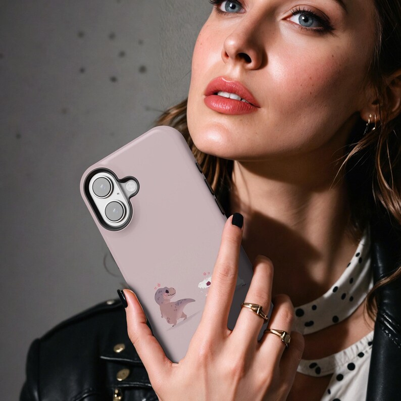 Puede incluir: Funda de tel&eacute;fono rosa claro con un dise&ntilde;o de dinosaurio y cupcake de dibujos animados. La funda tiene un recorte negro para la c&aacute;mara. El fondo es gris borroso. La funda est&aacute; siendo sostenida.