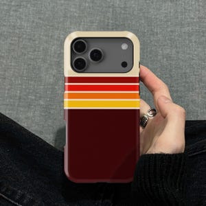 Puede incluir: Una funda para smartphone con un diseño retro. La funda presenta un borde color crema y una base de color rojo oscuro. Rayas horizontales en rojo, naranja y amarillo adornan la parte superior. Las lentes de la cámara del teléfono son visibles.