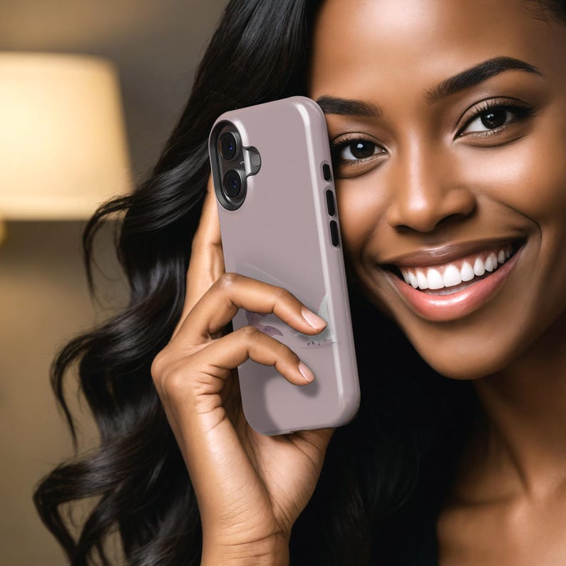 Puede incluir: Una funda de tel&eacute;fono de color malva claro con una c&aacute;mara de doble lente y botones laterales. La funda se sostiene en una mano con cabello largo y oscuro en el fondo. La funda del tel&eacute;fono es de color s&oacute;lido con un acabado liso.
