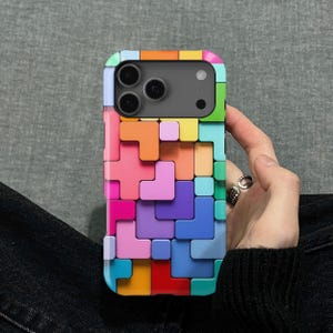 May include: A multi-coloured phone case with a Tetris block pattern. The case is composed of various shapes in shades of red, orange, yellow, green, blue, and purple. The phone case has a black camera module.