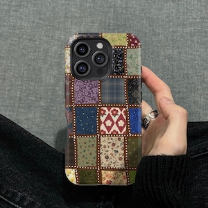 以下が含まれることがあります： 緑、紫、青、赤の色合いの様々な生地パターンが特徴の、パッチワークスタイルの携帯電話ケース。ケースは茶色の縁取りと、縁の周りに点線模様があります。携帯電話ケースは人が持っています。