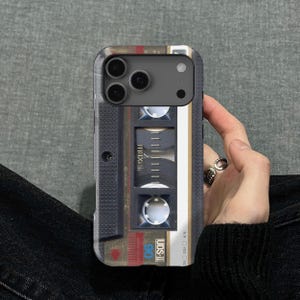 May include: A phone case with a vintage cassette tape design. The case has a black border and a detailed graphic of a cassette tape with the text "UDS-II 90". The case is held by a hand.