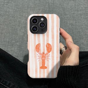 May include: A white phone case with vertical peach stripes and a watercolor illustration of an orange lobster. The phone case is held by a person wearing a black sweater and a silver ring.