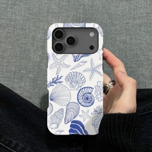 Puede incluir: Funda de teléfono blanca con un patrón de conchas marinas azules. El diseño presenta varias conchas, estrellas de mar e hipocampos. La funda se sostiene en una mano, mostrando la detallada ilustración náutica.