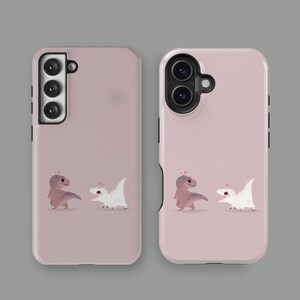 Puede incluir: Dos fundas para tel&eacute;fono de color rosa empolvado. Cada funda presenta una ilustraci&oacute;n de dibujos animados de un Tyrannosaurus Rex marr&oacute;n y un dinosaurio blanco. Las fundas est&aacute;n dise&ntilde;adas para diferentes modelos de tel&eacute;fono.