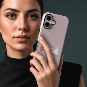 Puede incluir: Funda de tel&eacute;fono rosa claro con una ilustraci&oacute;n de dibujos animados de dos peque&ntilde;as criaturas parecidas a dinosaurios. Una es rosa y la otra blanca. La funda tiene un borde negro para la lente de la c&aacute;mara y la sostiene una persona.