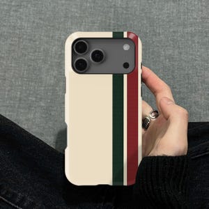 Peut inclure: Coque de téléphone crème avec un design à rayures verticales vertes foncées et bordeaux. L'étui a une découpe pour l'appareil photo et est tenu par une personne.