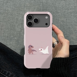 Puede incluir: Funda de tel&eacute;fono rosa claro con una ilustraci&oacute;n de dibujos animados de dos dinosaurios. Uno es de color p&uacute;rpura claro, el otro blanco. La funda tiene un parachoques protector y un recorte para la c&aacute;mara.
