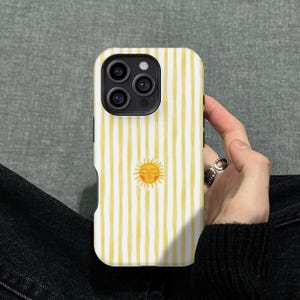 Może przedstawiać: Etui na smartfona z białym i żółtym wzorem w paski oraz grafiką słońca. Etui jest przeznaczone do ochrony telefonu. Etui jest trzymane w dłoni osoby.