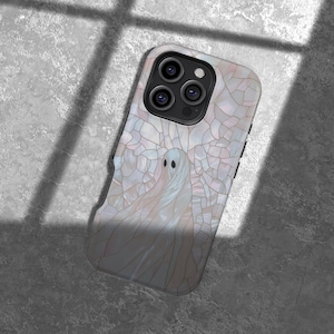 May include: A phone case featuring a ghost design. The case has a pearlescent, mosaic-like background with a white ghost illustration. The phone case has a black camera module and buttons on the side. The case is on a gray surface.