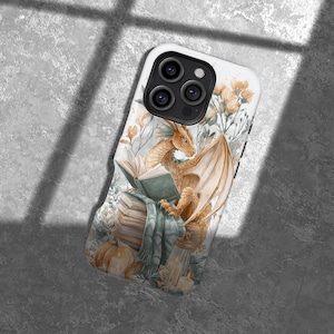 Op de afbeelding: Witte telefoonhoes met een aquarel illustratie van een draak die een boek leest. De draak is goudbruin met grote vleugels, zittend op een stapel boeken. De achtergrond bevat pompoenen, bloemen en een blauwe deken.