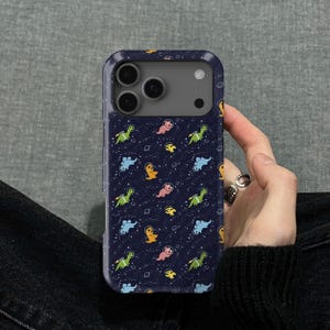 May include: A navy blue phone case featuring a repeating pattern of colorful cartoon dinosaurs in space suits against a starry background. The case has a protective bumper design and cutouts for the camera.